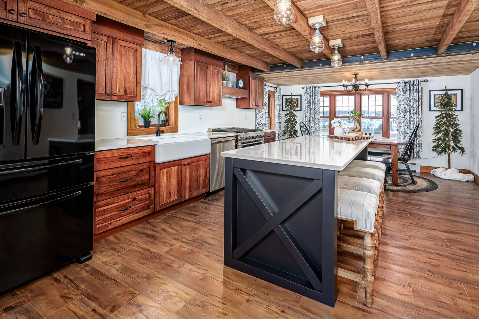Noah F. Boyle Cabinets Elizabethtown, PA Modern farmhouse kitchen with wood cabinets, black appliances, a white farmhouse sink, marble island with stools, wooden floors, and visible ceiling beams. Dining area in the background.