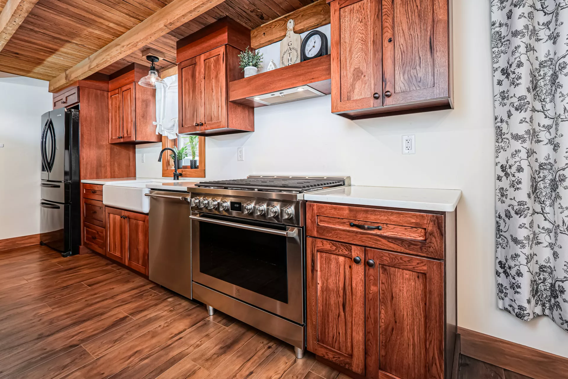 Noah F. Boyle Cabinets Elizabethtown, PA Modern kitchen with wooden cabinets, stainless steel appliances, a farmhouse sink, and wood flooring. A window with a white curtain and floral pattern is visible on the right.